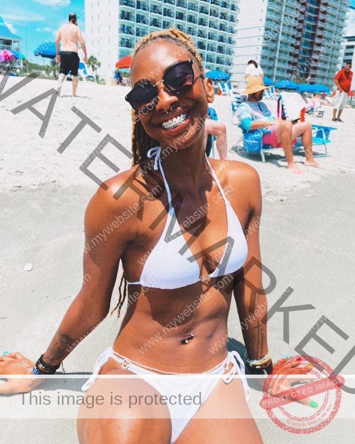 Serena Harrison sits on the beach in a white bikini, sunglasses and a big smile.