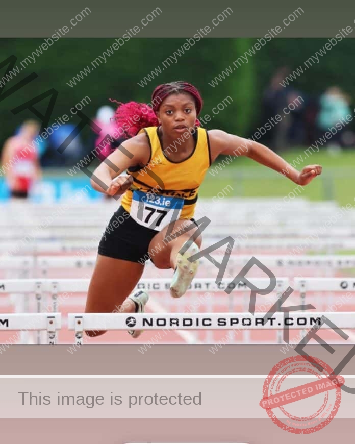 Okwu Backari wearing a yellow top and black shorts and number 77, clears a hurdle in a race.