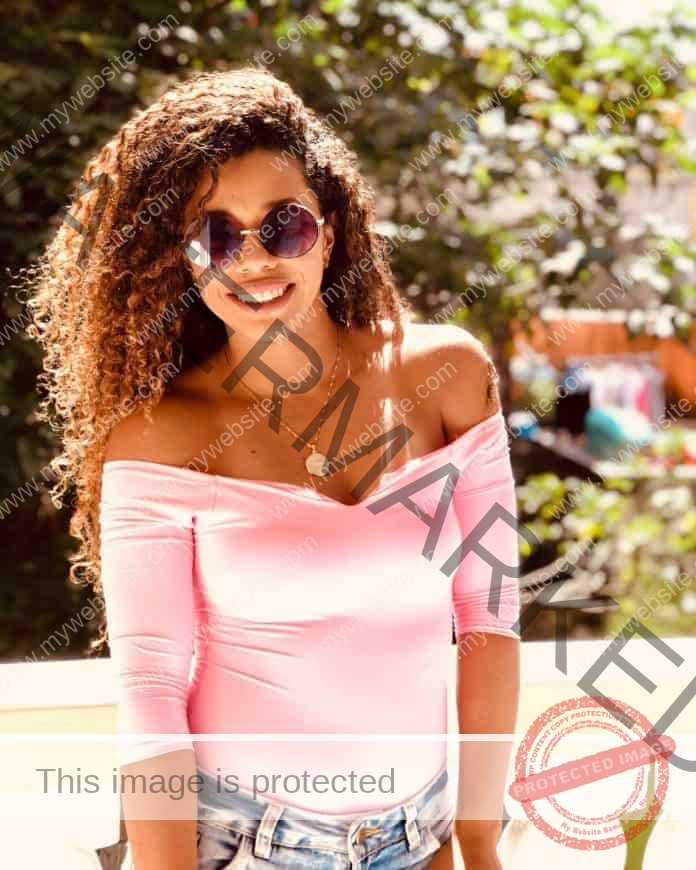 Andressa Fidelis standing in the sunlight, wearing a cute pink top, jeans and sunglasses.