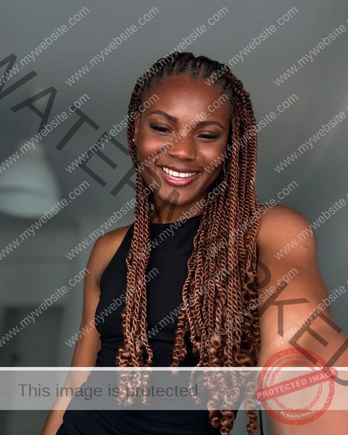 Agate De Sousa smiling for a selfie in a bedroom, wearing long braids and a black shirt - with a smile.