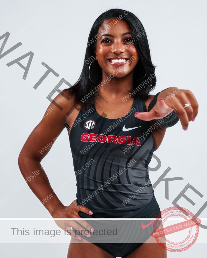Skylynn Townsend posing for a photo shoot in front of a white background, wearing her track uniform and pointing at the camera, smiling.