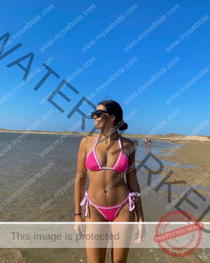Aitana Moscard standing on the beach in sunglasses, looking off into the distance, with a pink and white string bikini.