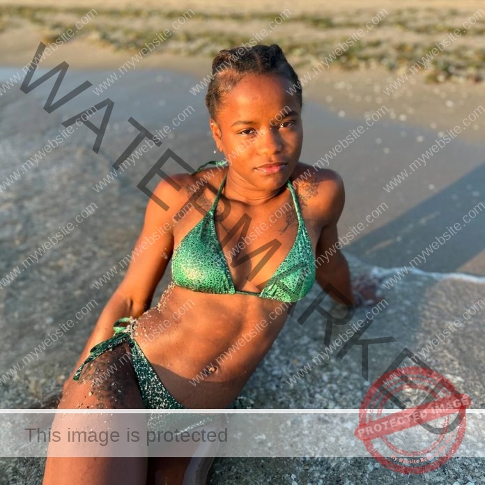 Alejandra M. Ferrari lying in the surf of the beach on her side, propped up on one arm, wearing a shiny green string bikini.