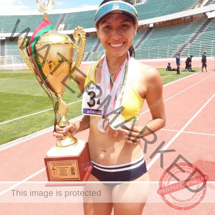 Lorenz Rios Paz, a smiling female athlete from Bolivia, stands on a track with a large trophy and medals; stadium seats behind.