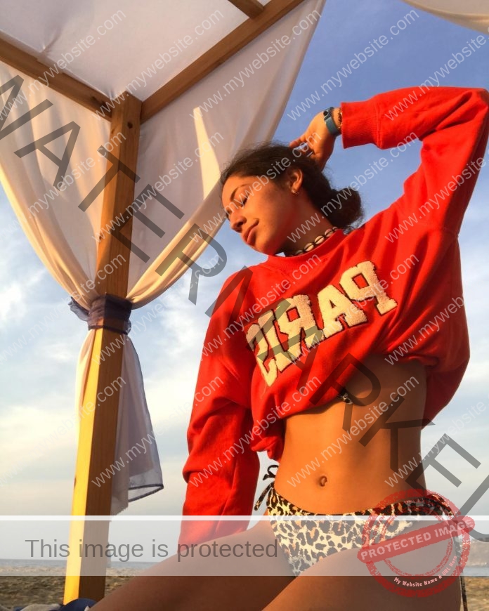 Sofia Delgado Yepez posing for a photo in the sun, under a fabric, wearing a red sweatshirt with Paris written on it, arm raised above her head.