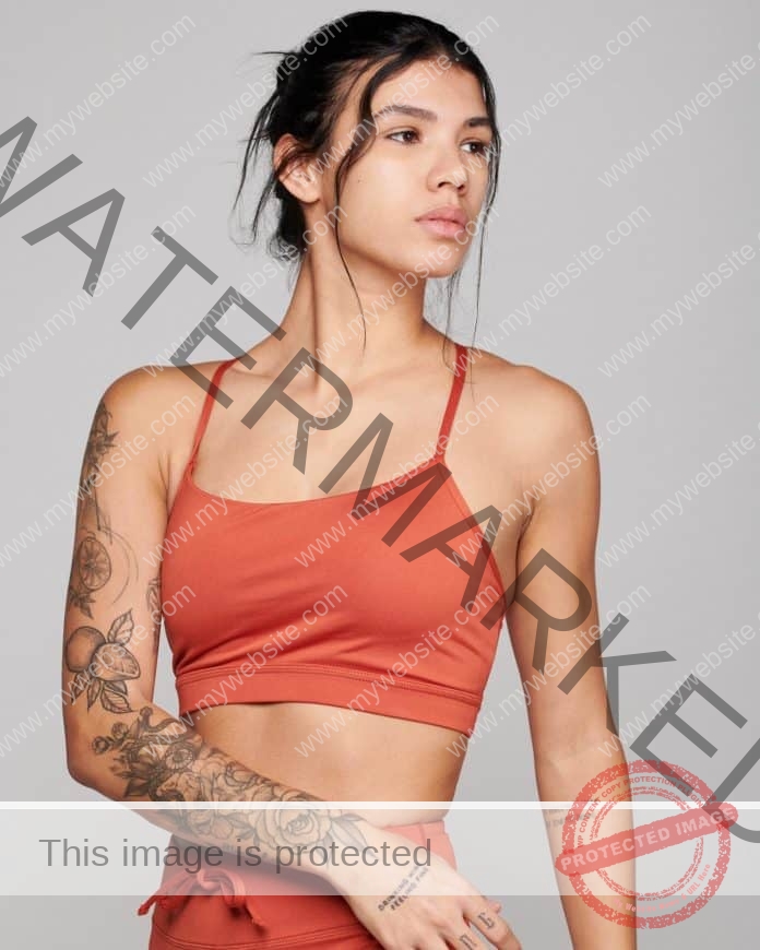 Paola Sarabia posing in front of a white background, wearing an orange tank top and pants, turning at the waist and showing her tattoos.