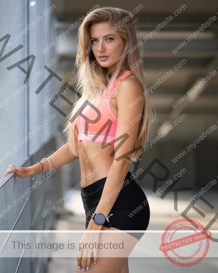 Alicia Schmidt standing facing a window, hand on a rail, wearing a pink sports bra and black short tights, looking at the camera.