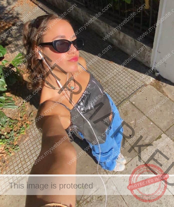 Isabella Conde de Frankenberg staking aa selfie, arm extended, wearing sunglasses and a black shirt and blue pants.