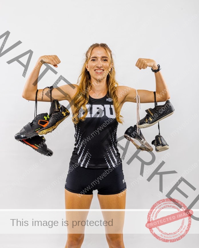Rebecca Pecora posing in her college track uniform against a white background, pairs if shoes draped over her flexed arms by the laces.