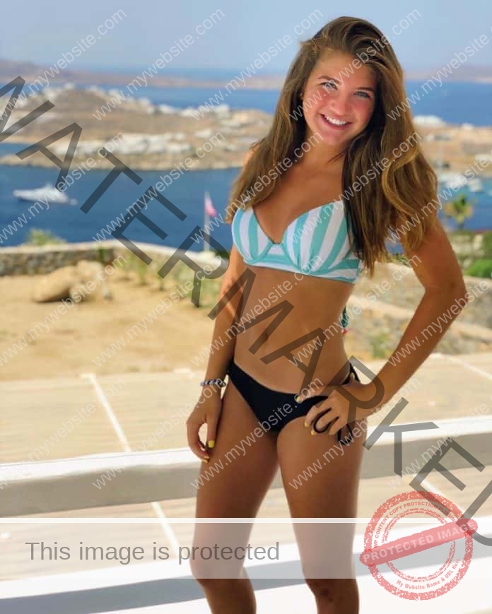 Allison Spalding standing on a blacony in front of the ocean, smiling and wearing a blue and white striped bikini top and black bikini bottoms.