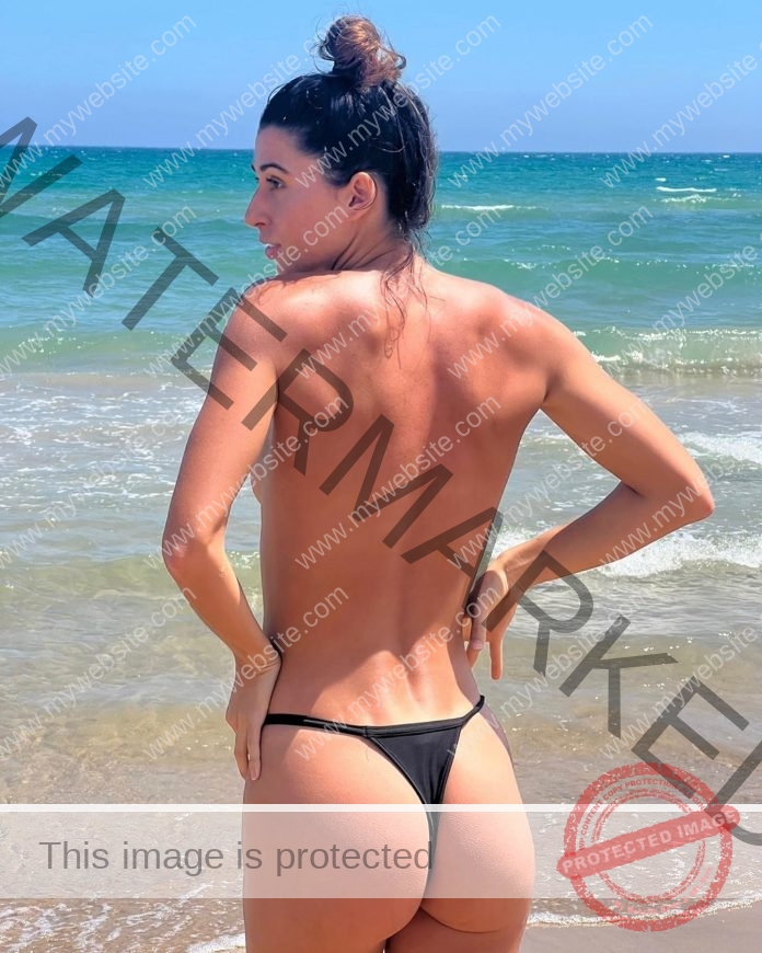 Sheila Prados stands on a sandy beach in Spain, facing the sea in a black bikini bottom, ocean and sky stretching behind her.