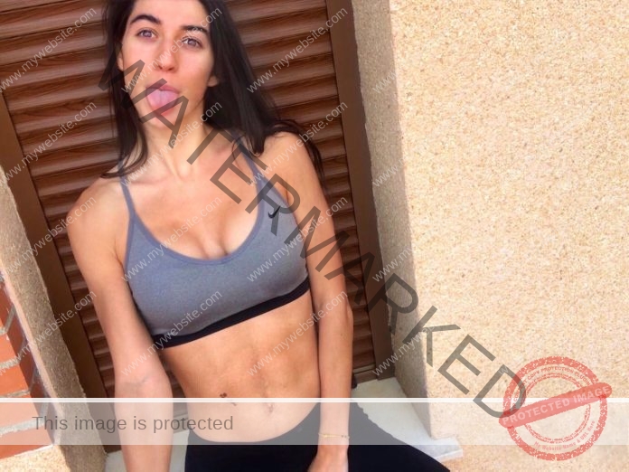 Sheila Prados, a track and field athlete from Spain, poses outside against a tan wall in gray sports bra and black leggings, her long dark hair cascading as she looks slightly upward with a relaxed expression.