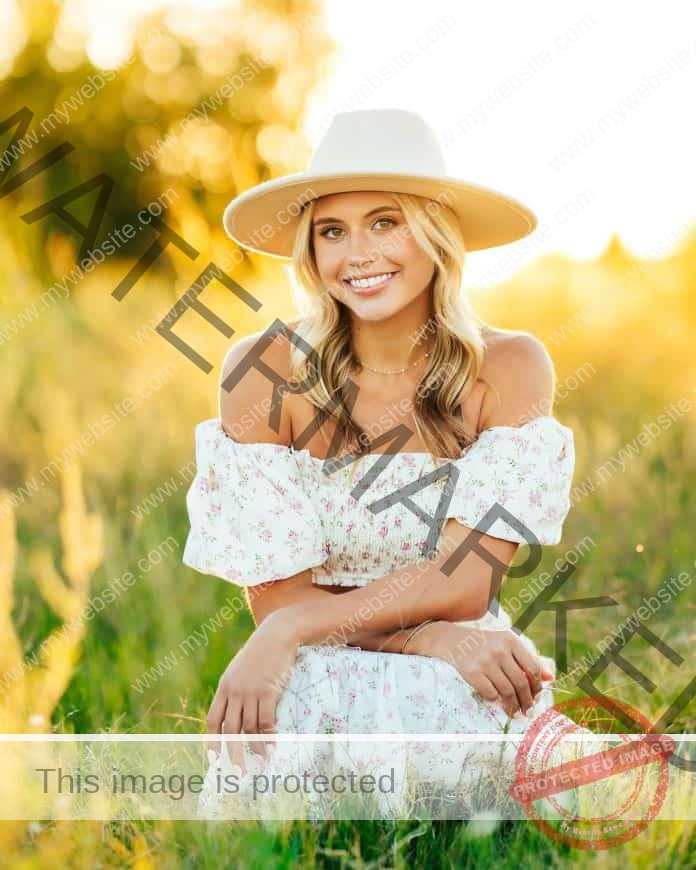 Ava Goetz posing for a professional photo shoot in a tall grass field, wearing a country dress and cute straw hat.
