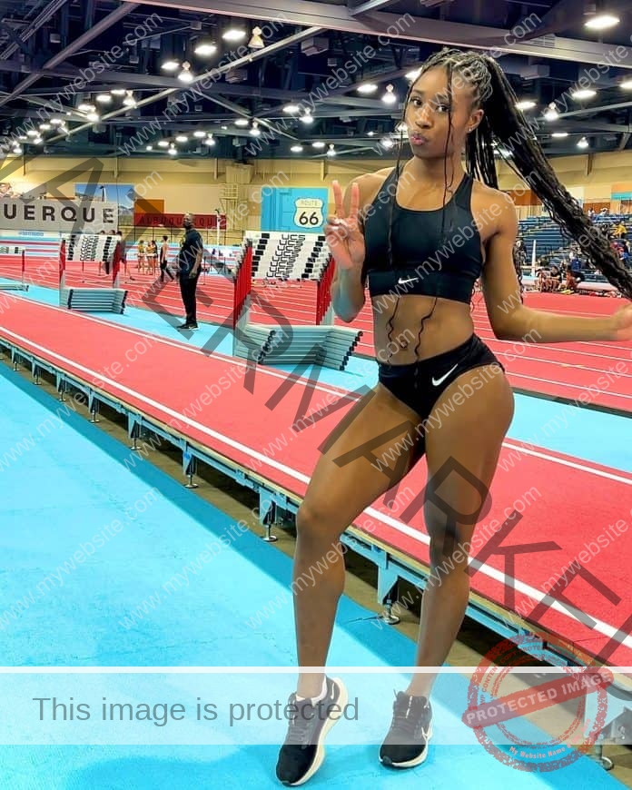 Aniella Delafosse standing trackside, posing for a picture in her track uniform, right leg cocked and braids in her hands.