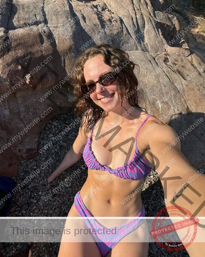 Anna Grimaldi taking a selfie in the ater by rocks, wearing a small purple string bikini, sunglasses and a smile.