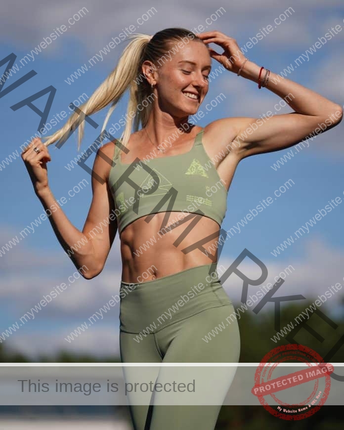 Anna Ryzhykova stands outdoors in a light green sports bra and leggings, smiling confidently, holding her ponytail under a blue sky.