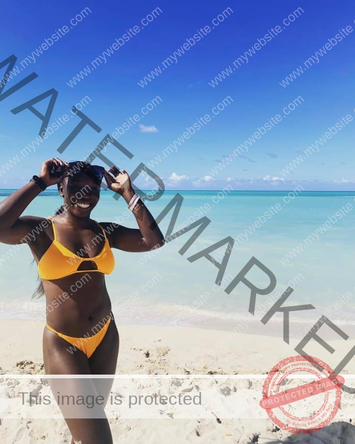 Dahlia Barnes standing on the ebach wearing a yellow bikini, hands on top of her head holding her sunglasses.