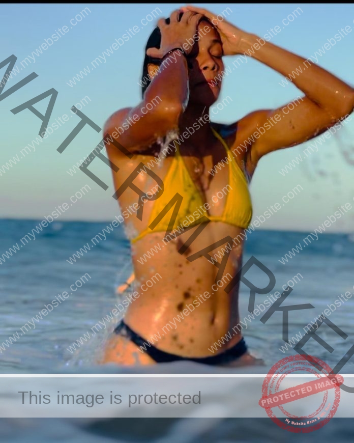 Daniela Alejandro waist deep in the ocean, slicking back her wet hair and wearing a yellow bikini top with black bottoms.