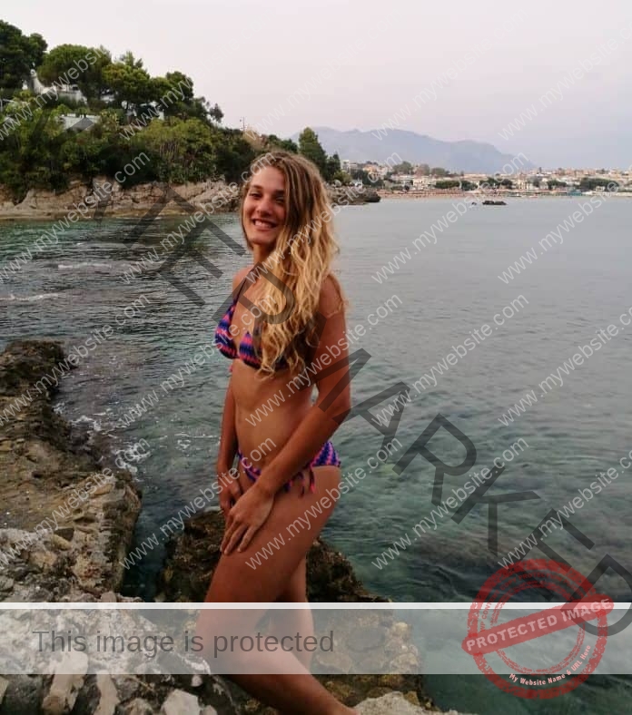 Elisa Tallini standing on the rocky beach, wearing a bikini.