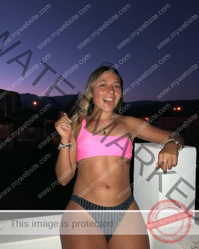Agustina Herrera smiles in a pink and black bikini outdoors at dusk, leaning on a white wall with mountains and lights behind her.