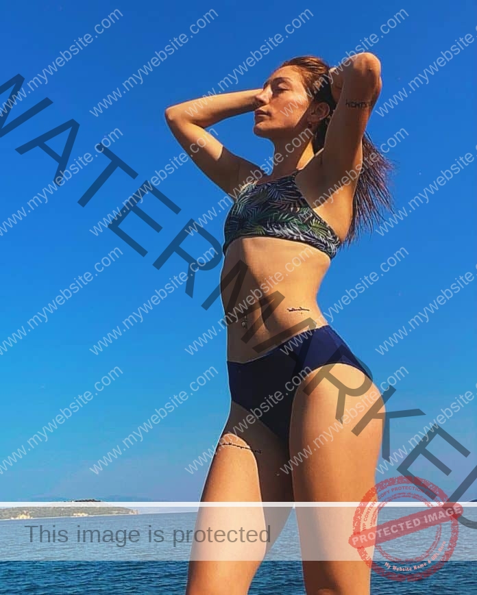 Beyzanur Yavuz standing by the oceaqn in a nice bikini, hands running through her hair as she stands sideways from the camera.
