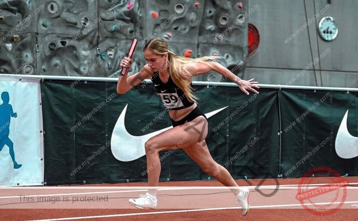 Lilijan Neerot sprints mid-stride on an indoor track in black sportswear, baton in hand, with a climbing wall and Nike logos behind.