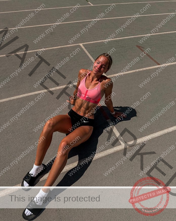 Lilijan Neerot in a pink sports bra, black shorts, and slide sandals sits smiling on a sunlit track and field, enjoying the day.
