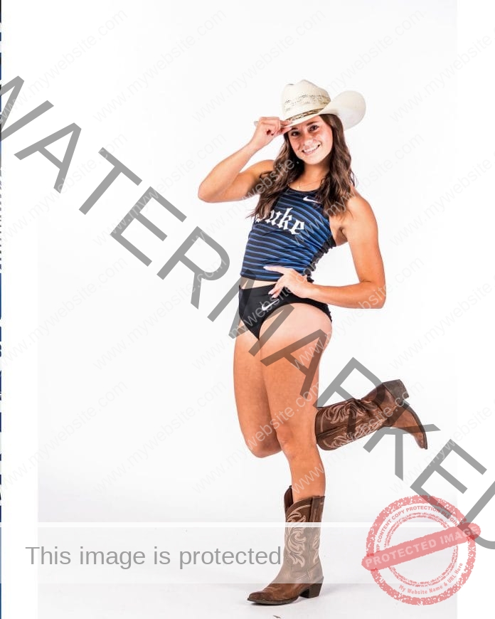 Valerie Jirak posing in front of a white bhackground, wearing her college track uniform, a cowboy hat and a big smile.