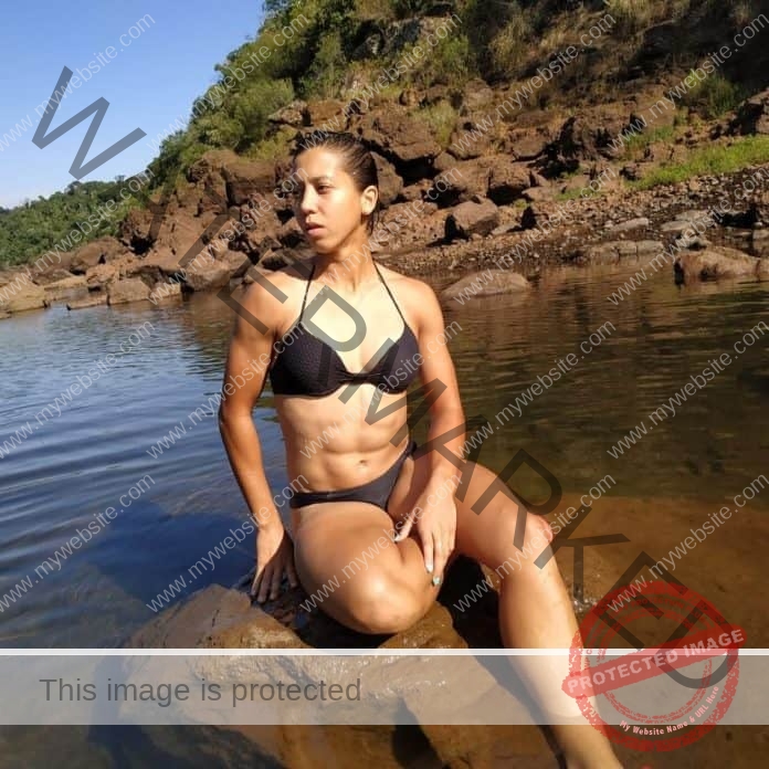 Vic Nieradka Violante sitting on a rock in the water, wearing a very small black string bikini to show off her ripped abs.