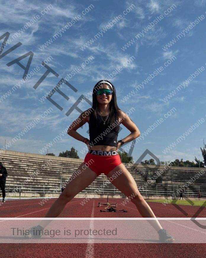Karla Miranda stands confidently on a track in sunglasses and sportswear, hands on hips, legs apart, with empty stadium bleachers behind.