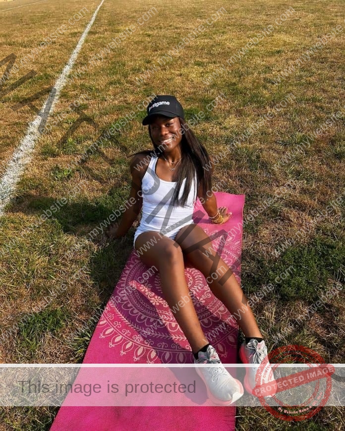 Manie Mevo sits on a yoga mat in the grass, wearing a white tank top, hat and shorts, smiling at the camera.
