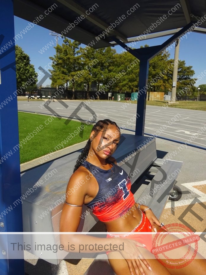 Sophia Beckmon sits on a metal bench in a red and navy USA uniform by a sunny track, shaded, with trees and fence behind.