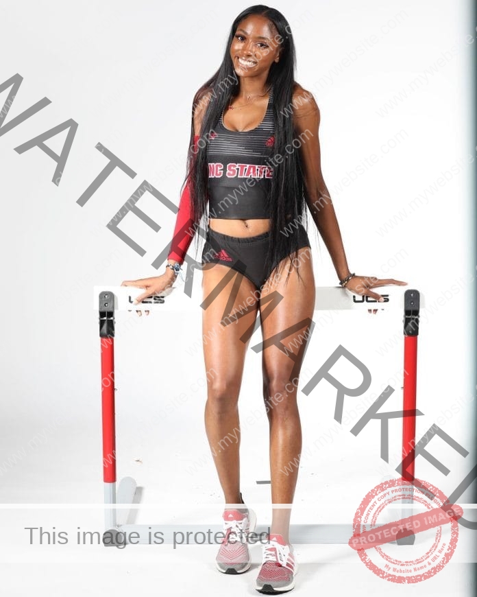 Timara Chapman posing in front of a white backghround for a photo shoot in her college track uniform, standing in front of a hurdle.