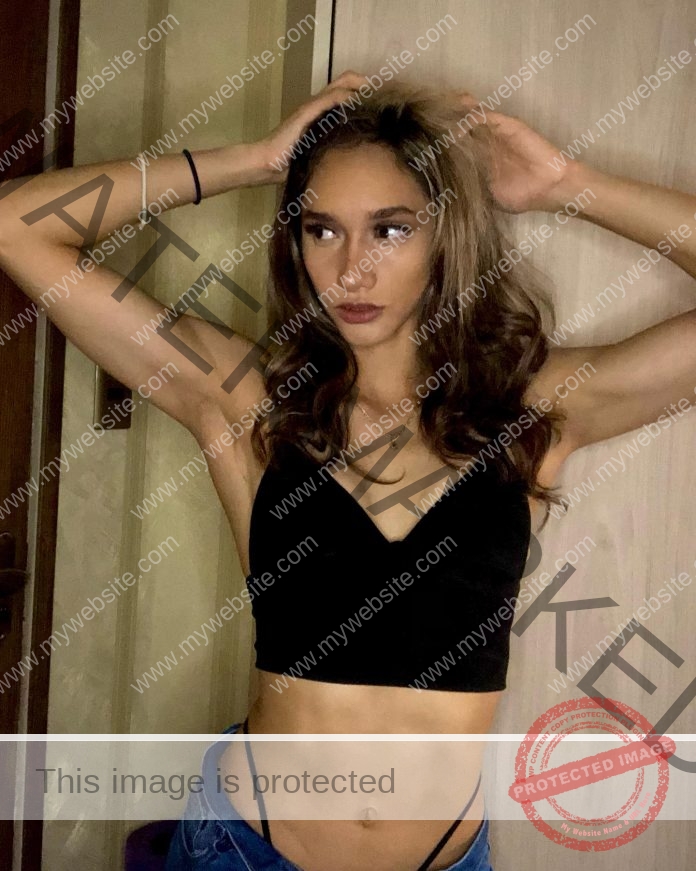 Leticia Arispe with long brown hair wears a black crop top, posing hands on head indoors by a wooden door. Watermarks are visible.