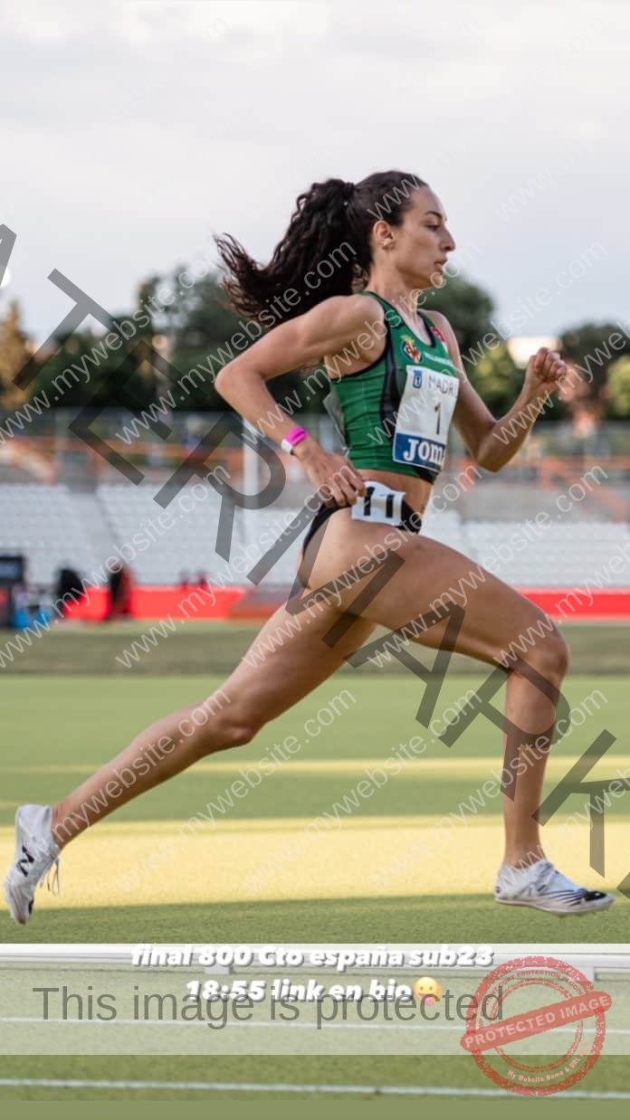 Yurena Hueso runs on the track in a green and black uniform during Spain's U23 800m final, with stadium seats and trees behind.