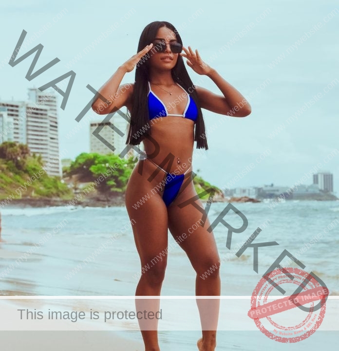 Karen Santana posing near the water in her blue string bikini and sunglasses.