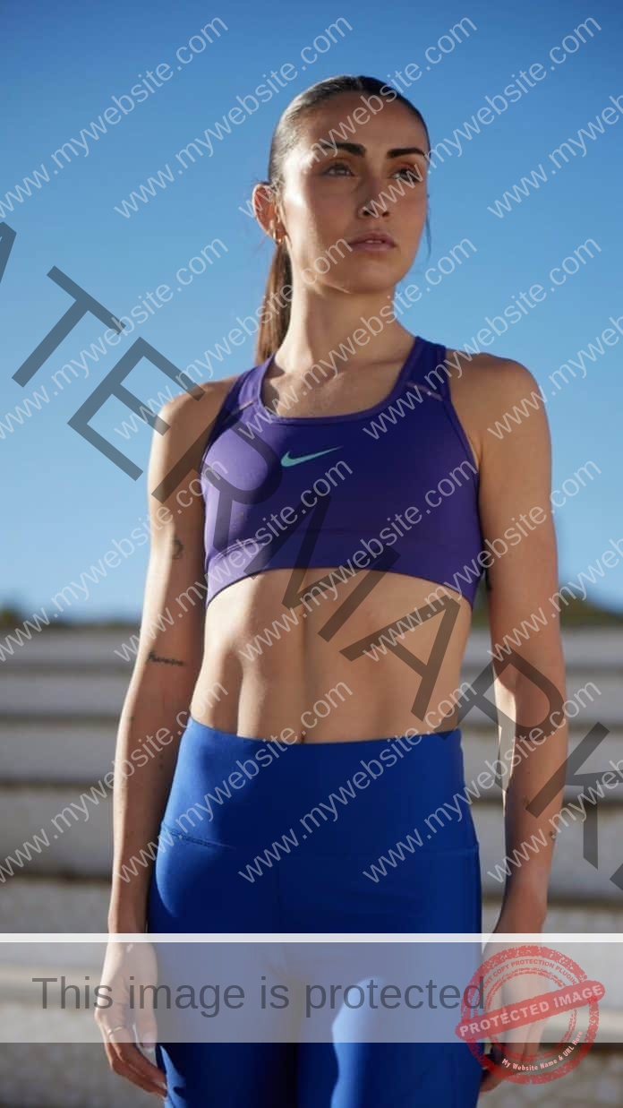 Marta Pla stands outdoors on stone steps in a purple sports bra and blue leggings, gazing determinedly into the distance under a blue sky.