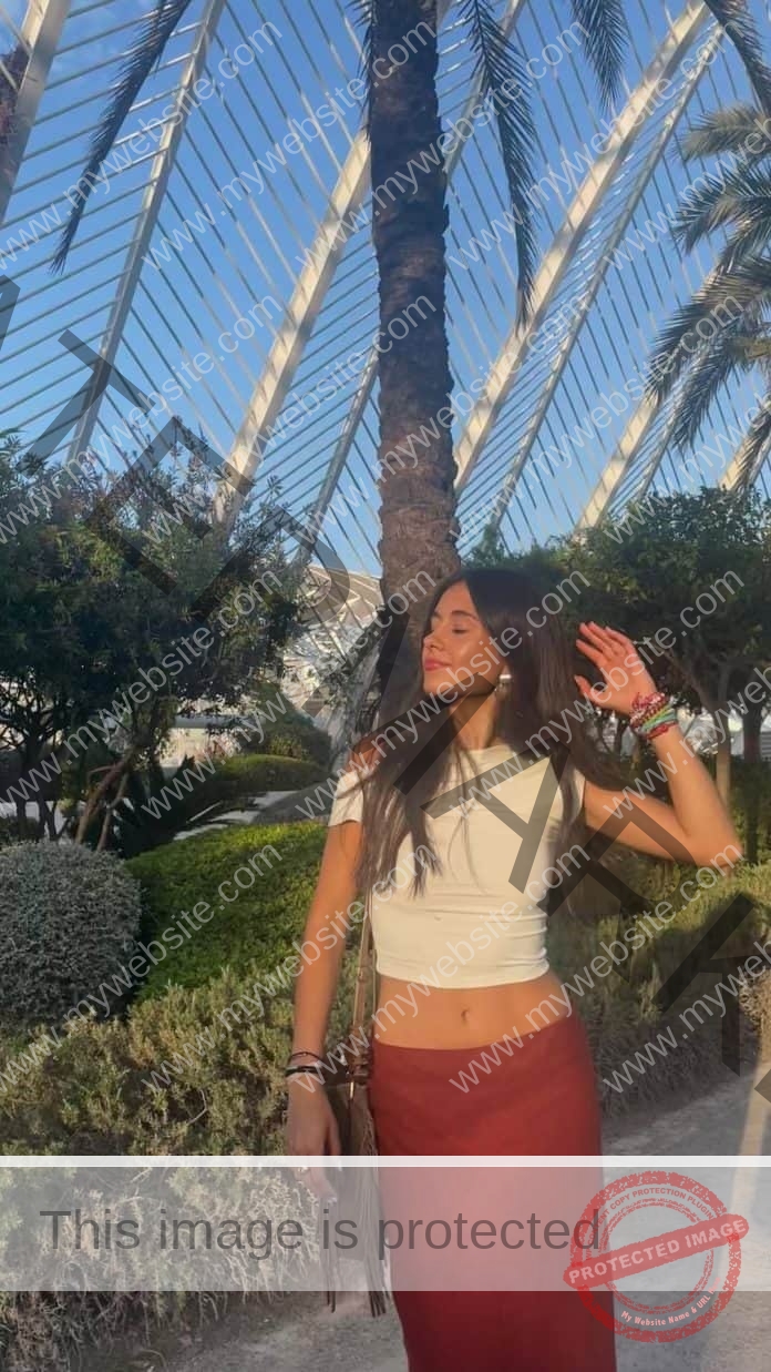 Celia Castro Padin standing in front of a telephone pole, wearing a white crop top and red pants, midriff showing, as she smiles and runs her fingers through her hair.