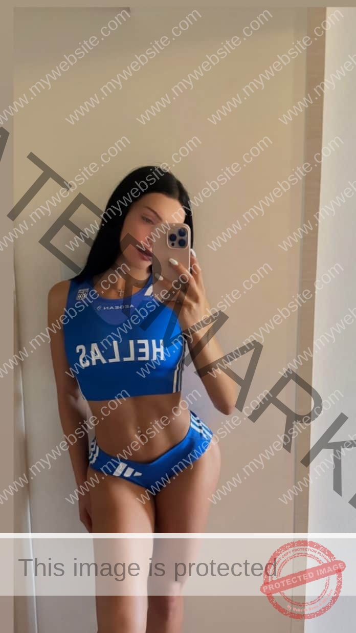 Kalliopi Remeplou with long dark hair takes a mirror selfie in a blue "HELLAS" sports outfit, relaxed against a beige wall.