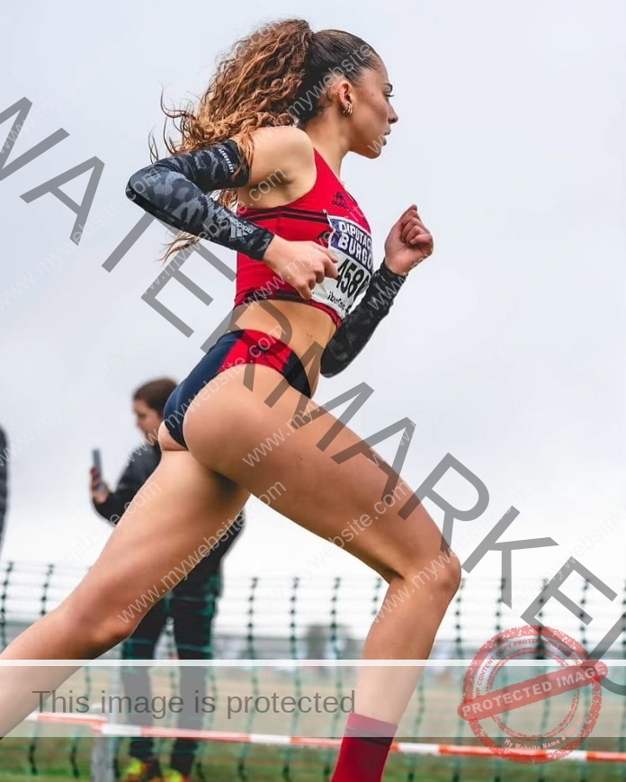 Amanda Roman, a female track athlete in a red crop top and navy shorts, runs mid-stride with a race bib outdoors in Spain.