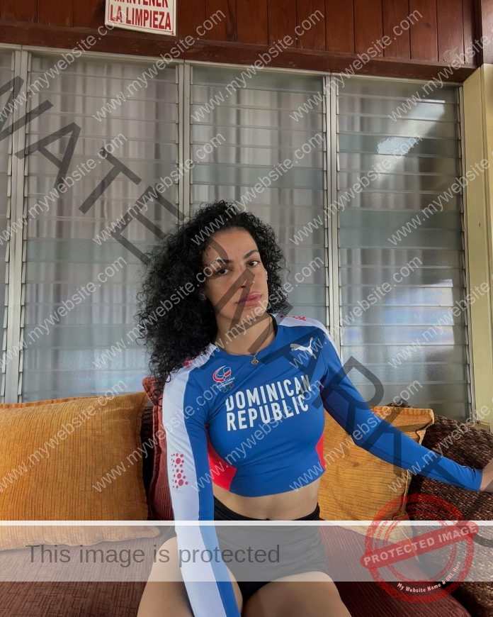 Bianca Acosta, with curly hair, sits on an orange couch in a blue Dominican Republic track and field shirt by a window with blinds.