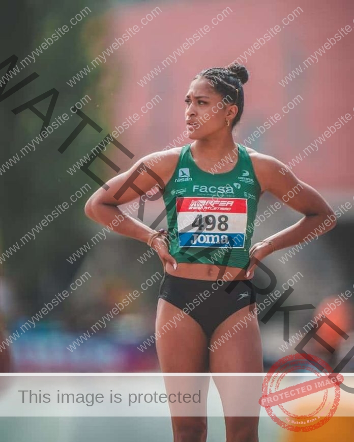 Irene Elewe, a focused female track athlete in green top and black shorts, bib 498, stands hands on hips at an outdoor event in Spain.