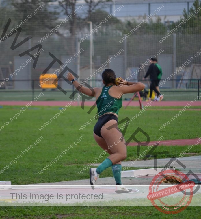 Eva Luna Luzano, a female Spanish track athlete in green sportswear, readies to throw shot put on an outdoor field with greenery.