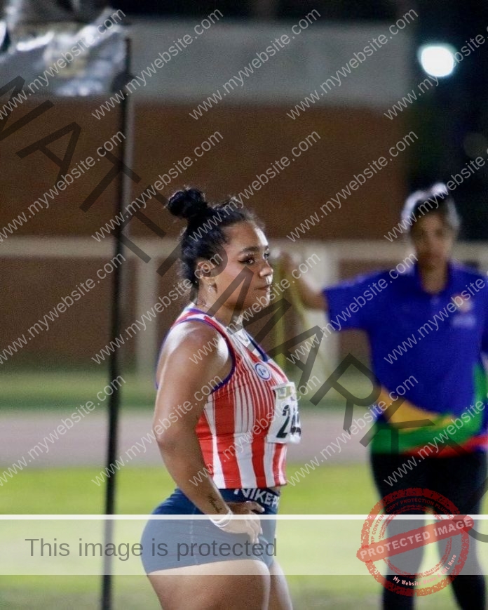 Fiora Veloso stands on the track in a red and white striped top and blue shorts, preparing to compete at night under sports lighting.