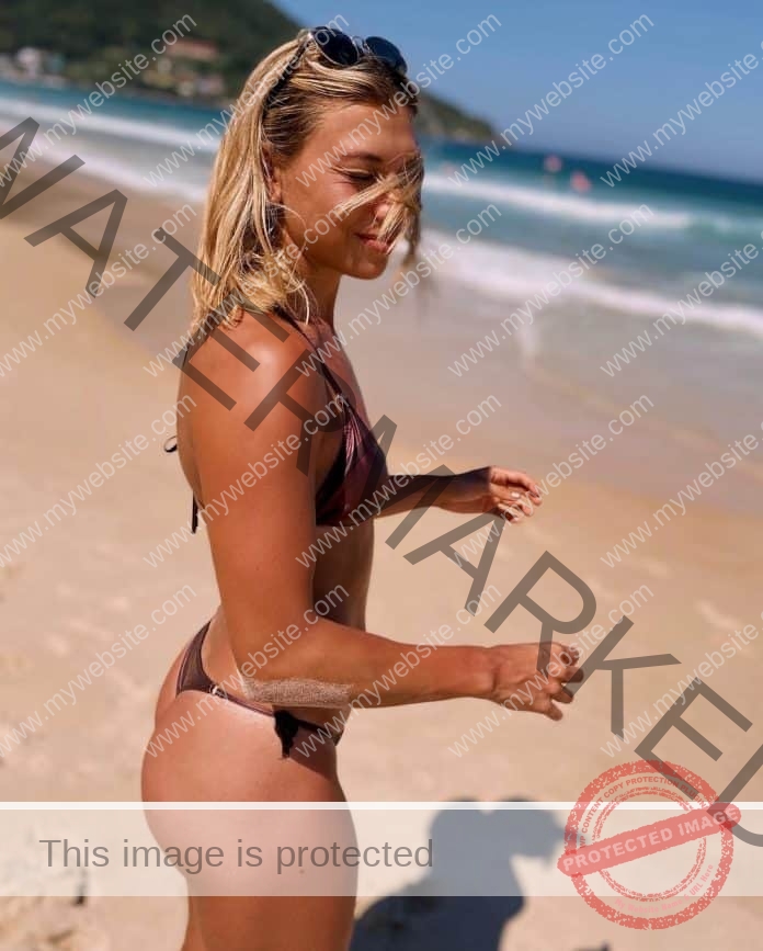 Guillermina Cossio standing on the beach in a string bikini, caught in a candid photo with a smile and hair in her face.