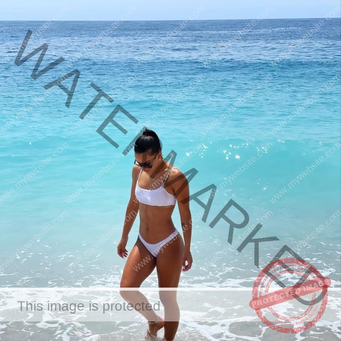 Iljana Beqiri strolls along the shoreline in a white bikini, clear blue water and gentle waves shimmering under a bright sky.