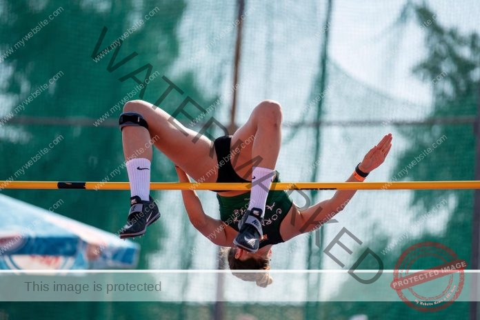 Irene Clemente, in sportswear and knee braces, arches mid-air over a high jump bar at a track event, green net and trees behind.