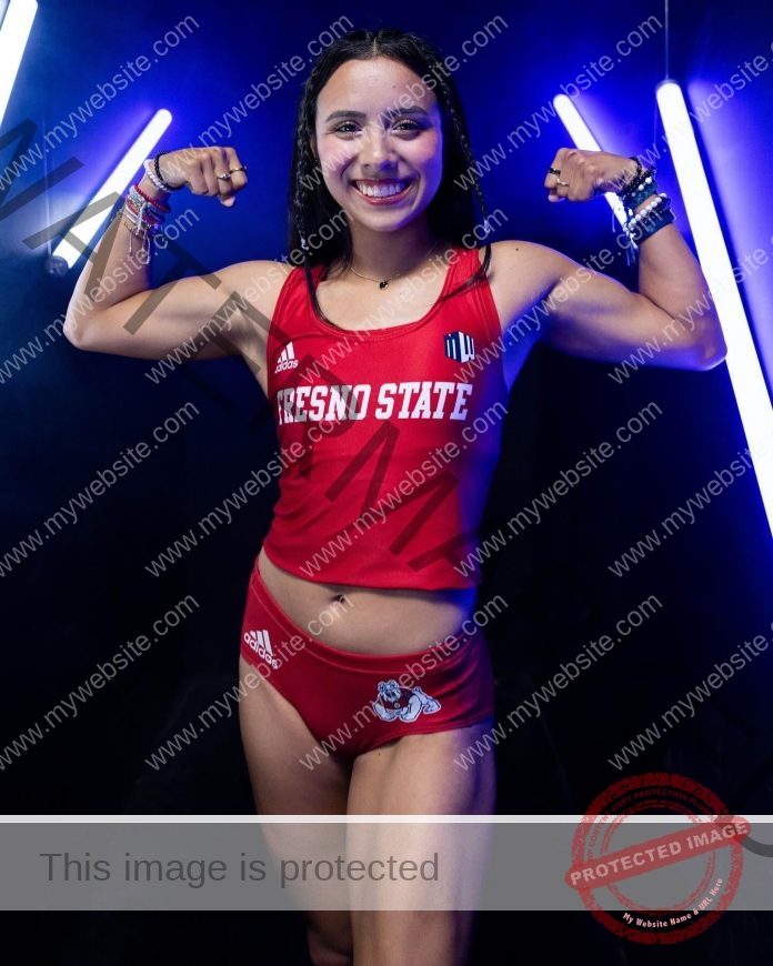 Itzia Mejia in a red Fresno State uniform flexes her arms and smiles before blue neon lights, bracelets on both wrists, representing Mexico.