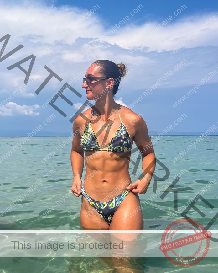 Janna Doumpas stands in clear, shallow ocean water on a sunny day, wearing a patterned bikini and sunglasses, with clouds behind.