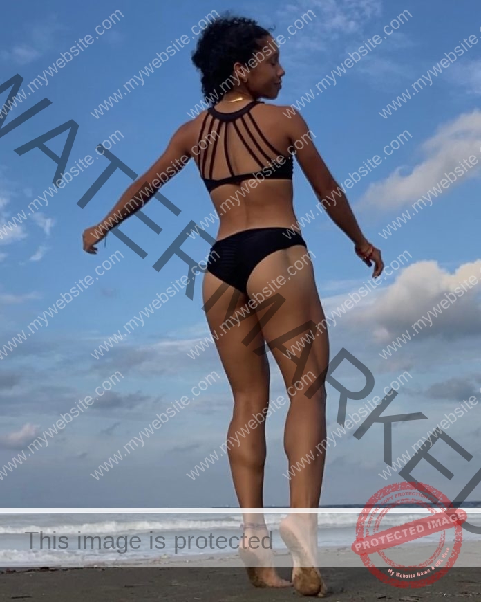 Jatziri Torales stands barefoot on sand in a black bikini, arms wide, looking at the ocean under blue sky—spirit of Mexico shining.